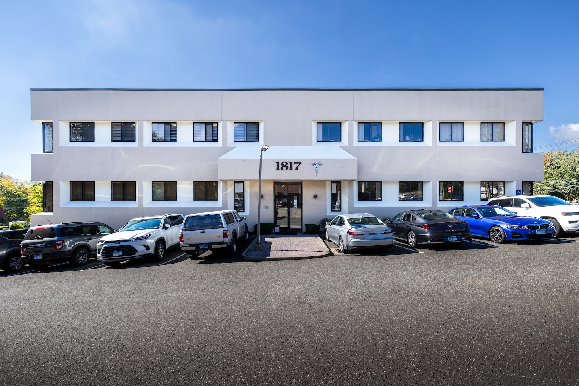 The image shows a multi-story building with a white facade, featuring a prominent entrance and a parking lot filled with various vehicles.