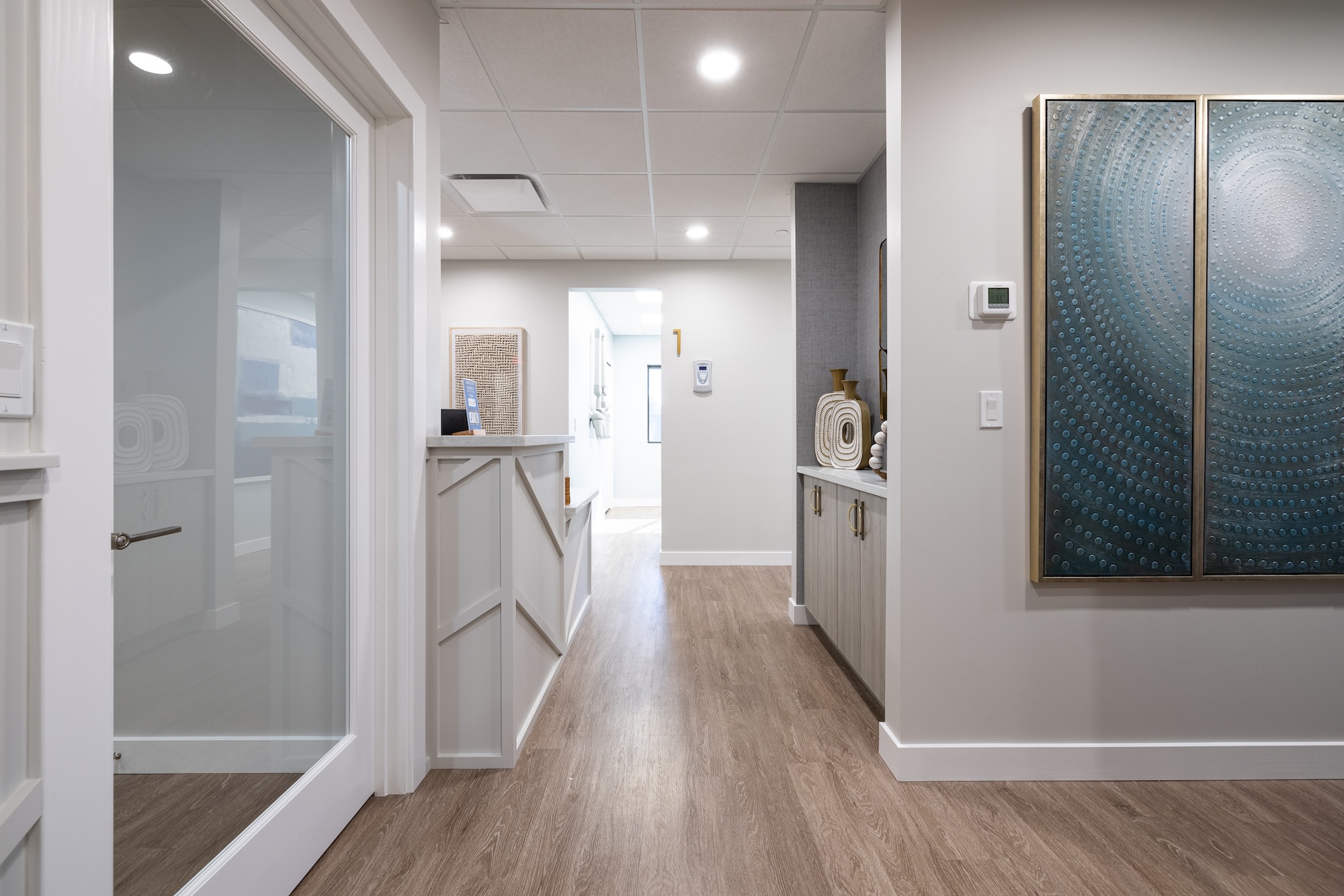 The image depicts a modern, well-lit hallway with a hardwood floor and white walls, leading to an open door revealing a kitchen area.