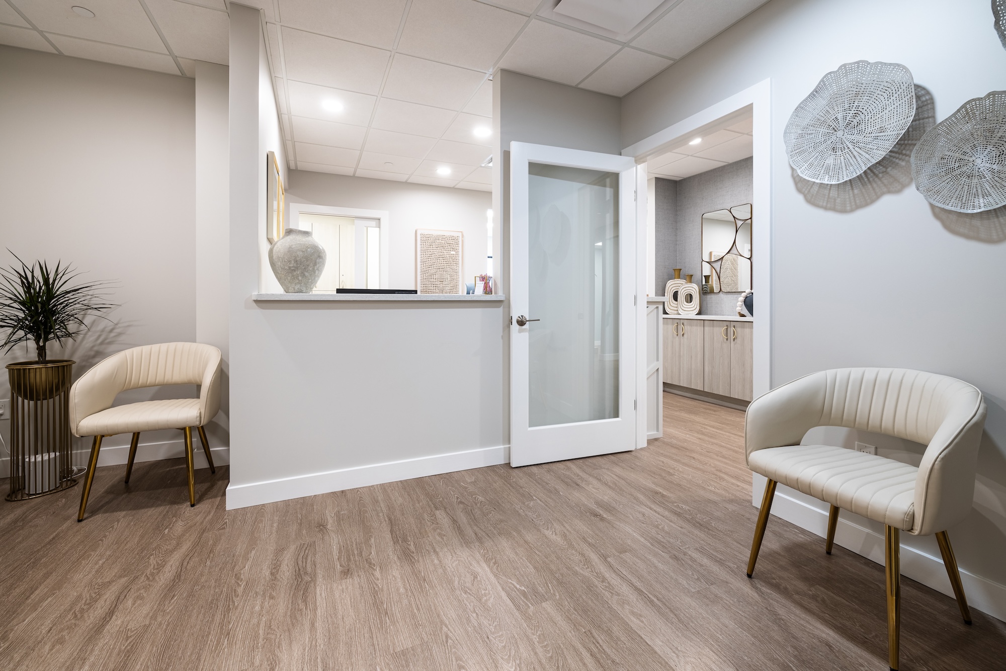 The image shows an interior space, likely a waiting or reception area, with a modern design featuring a large open floor plan, light-colored walls, a wooden floor, and minimalist furnishings.