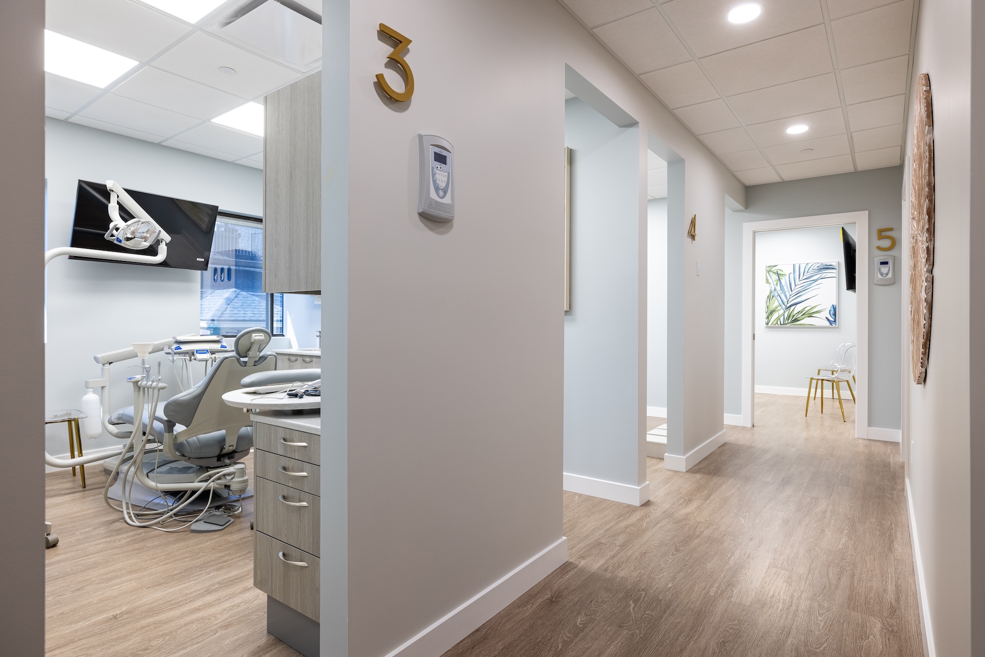 The image shows an interior space that appears to be a dental clinic, featuring a reception area with a desk and chair, a waiting room with chairs and a television screen displaying what looks like a nature scene, and a hallway leading to examination rooms.