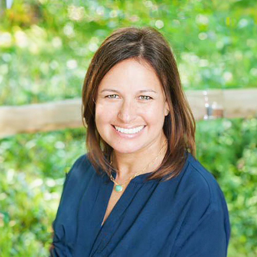 The image shows a woman with a smile, wearing a dark top and standing outdoors.