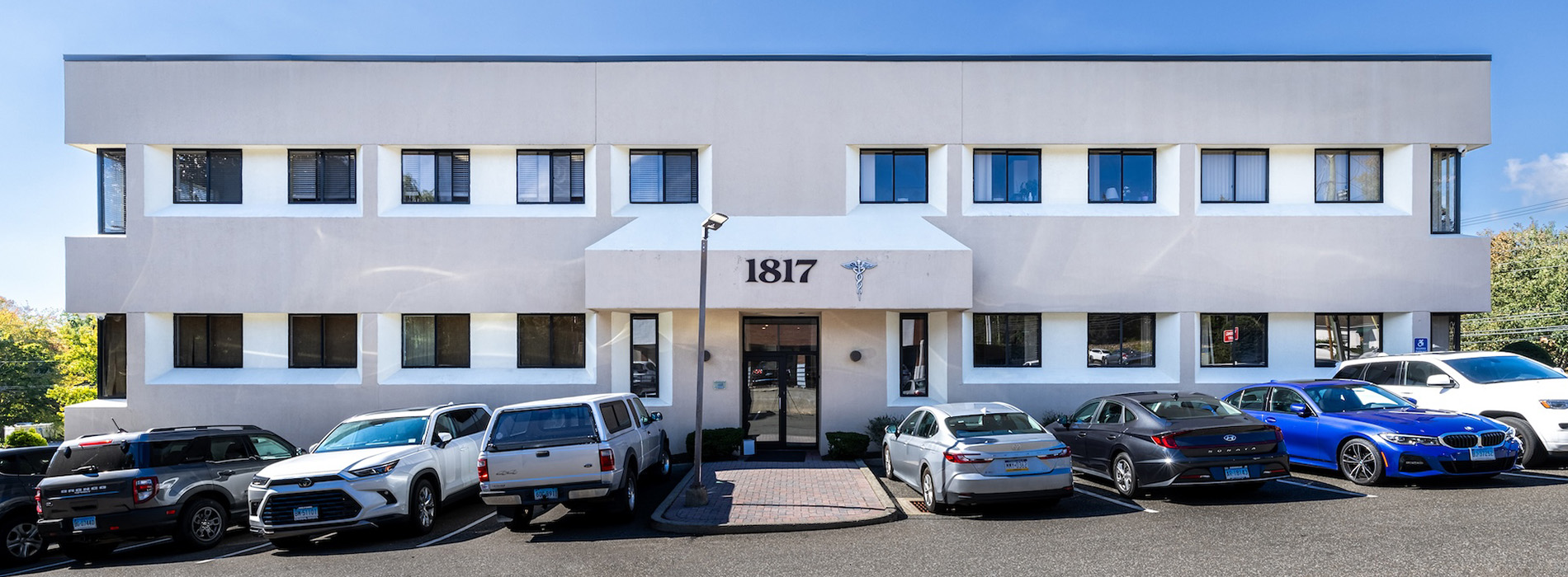 The image shows a modern, two-story building with a white exterior and multiple windows. It appears to be a commercial or office structure with a clear sky in the background.