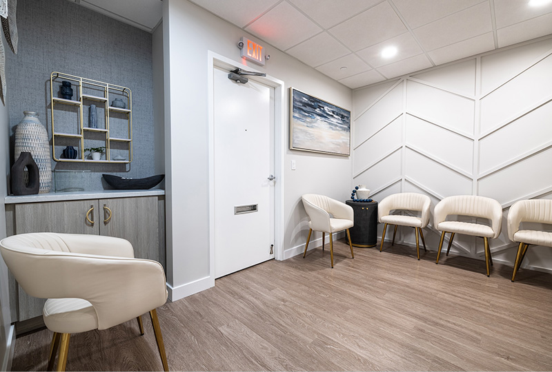 The image shows an interior space that appears to be a waiting room or reception area, featuring a row of chairs with a modern design, a door, and a portion of a wall-mounted artwork.
